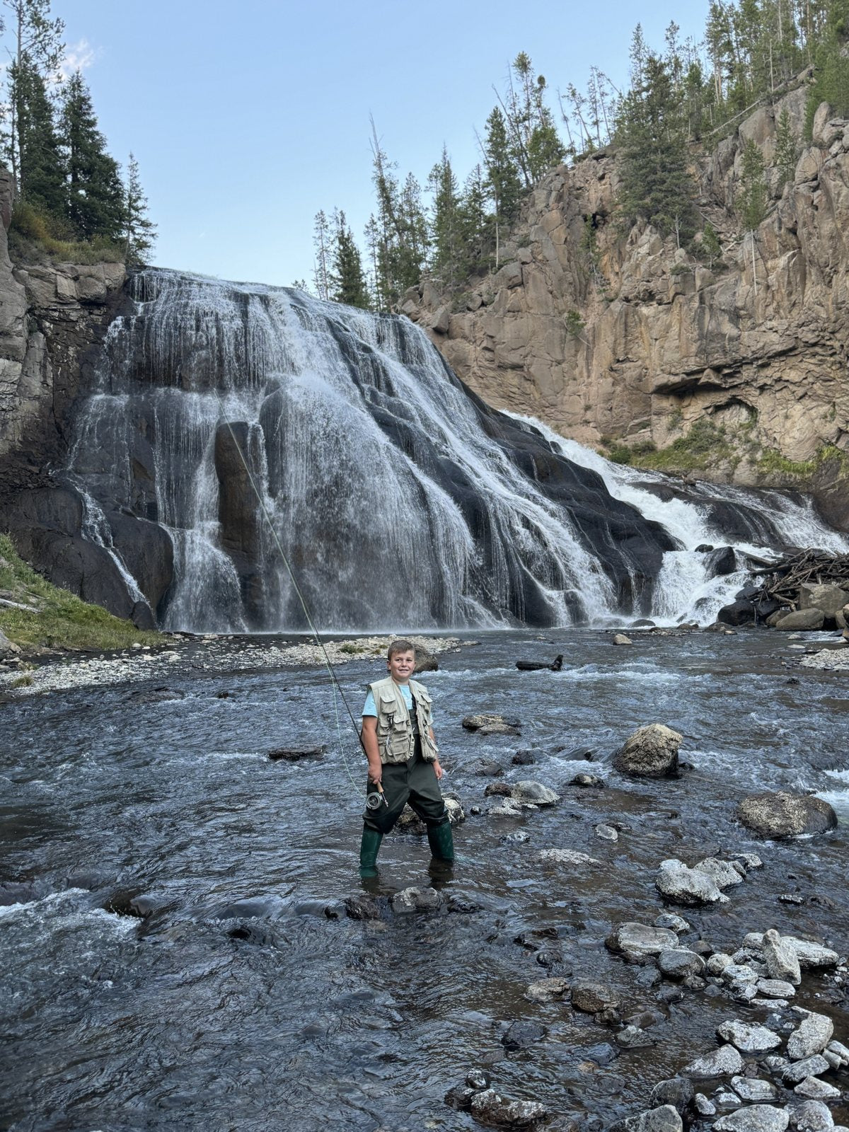 Caden wading a rocky river below a tall waterfall