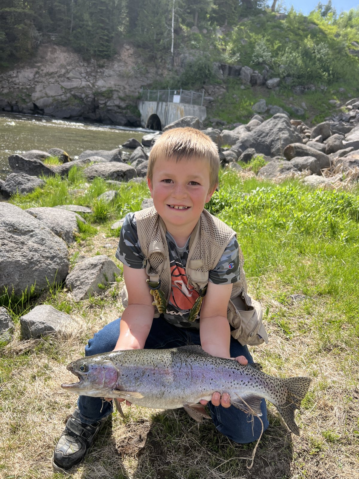 Young Caden kneeling on the riverbank holding a rainbow trout